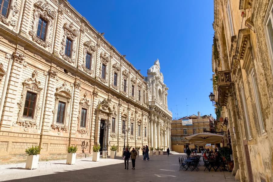 Groups of people walk between ornate golden stone buildings in Lecce.