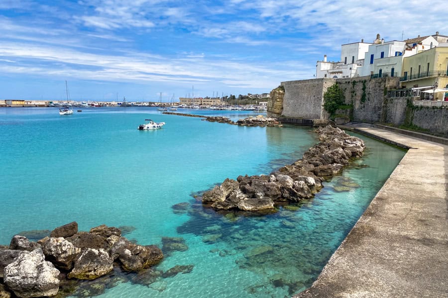 A harbour of crystal clear water with a boat on it, edged by a stone wall capped with buildings. 