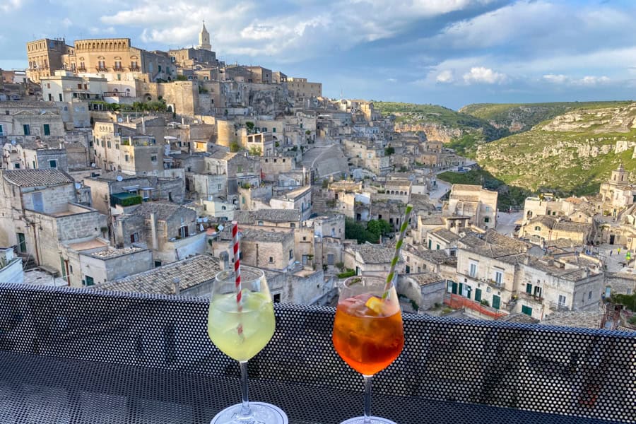 Two drinks on a table with the buildings of Matera rising in the background.