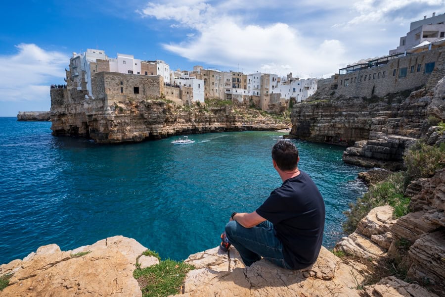 A man looks across water to buildings atop a cliff in Polignano a Mare, top stop on a Puglia trip.