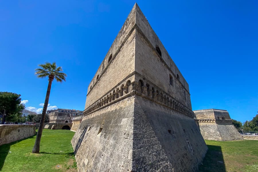 A stone castle rises from a grassy moat with a tall Palm tree, one of many fortresses you’ll see when you visit Puglia. 