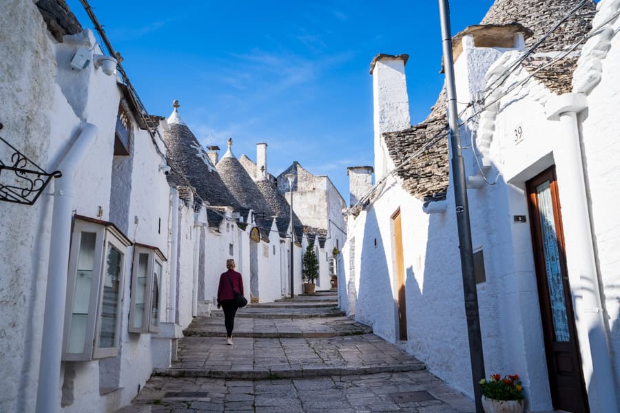 A woman walks along a cobbled lane between rows of white houses with pointed roofs on our itinerary for Puglia.