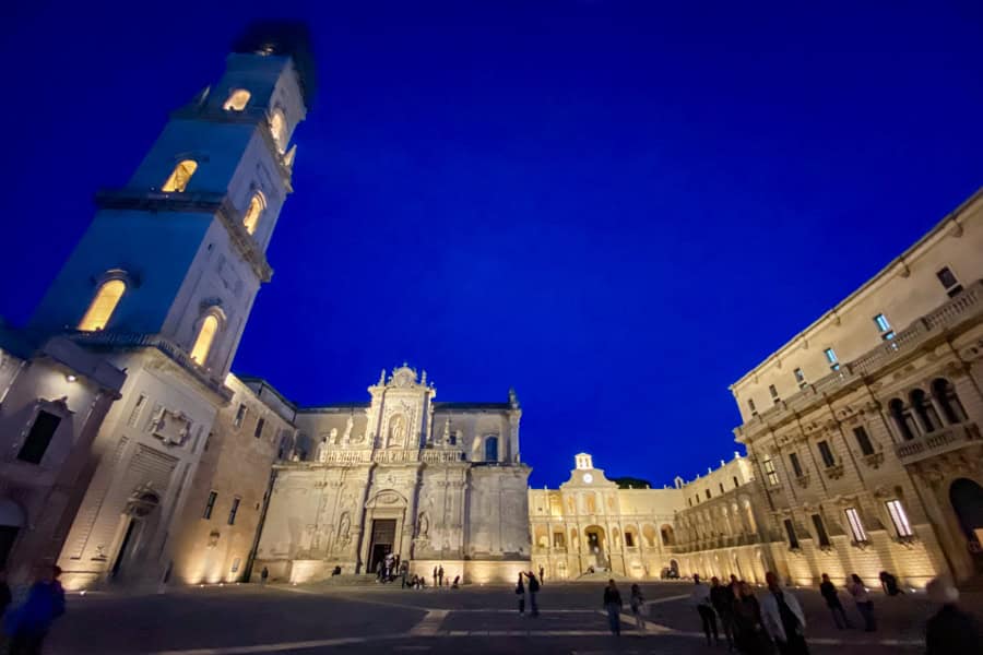 Lit up golden stone buildings edge a square in Lecce at night.